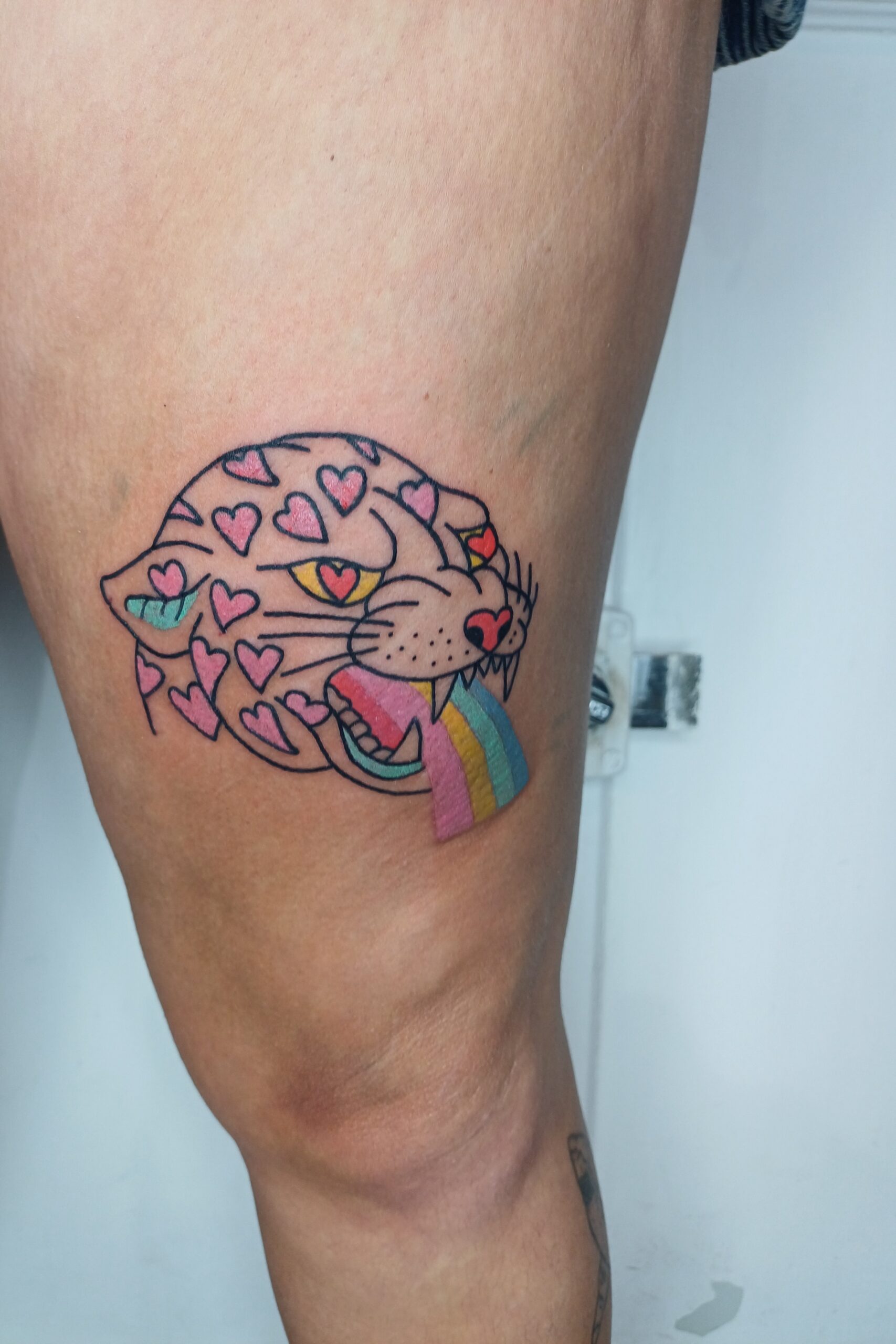 Foto van een het hoofd van een panter. De panter heeft een regenboog tong en hartjes op de kop. De tattoo werd op de dij gezet.
